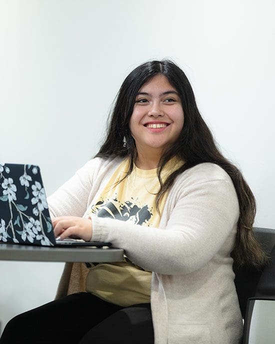 A student sits at a table. There is an open laptop on the table and their hands are resting on the keyboard.