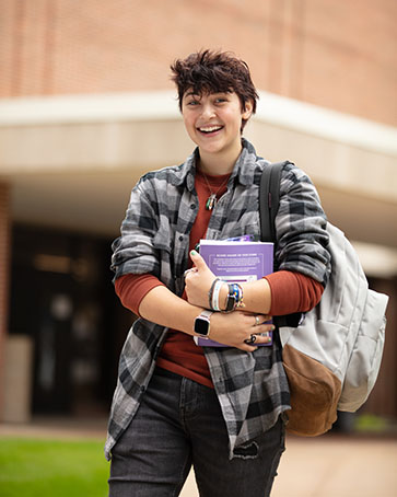 A student stands outside smiling. They are holding a book and have a backpack on one shoulder.