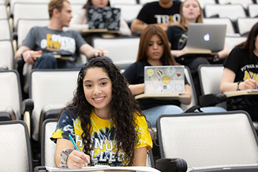 A student sits at a desk in classroom, there are other students sitting in the seats behind them.