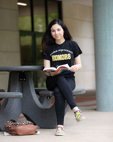 A student sits on a picnic table bench. She is wearing an "Honors College" t-shirt and is holding a book on her lap