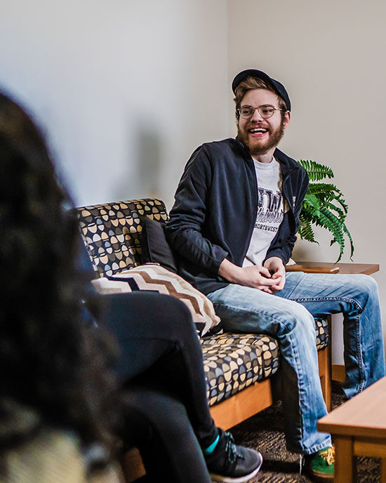 A PNW student sits on a couch and smiles at someone off screen.