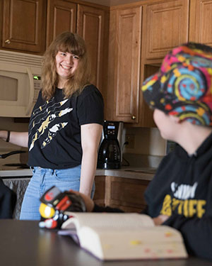 A student stands in the background smiling at another looking away from the camera, sitting at a table with a book
