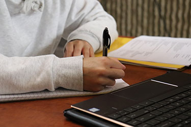 A person sits at a table with both arms resting on they table. They are holding a pen and writing on a piece of paper, there is also an open laptop on the table in front of them. The photo is focused on the person's arms and the table so that the person's face is not pictured.