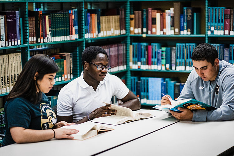 Three students sit at a table in the library. They are all reading books.