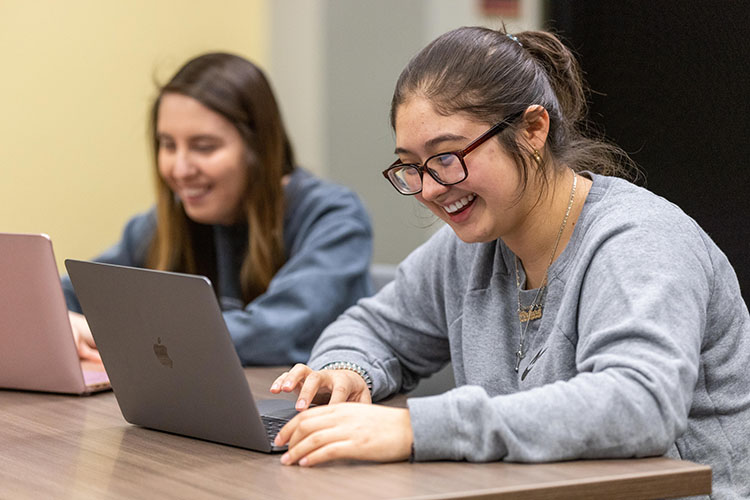 Two students sit at a table. They are both looking at laptops.