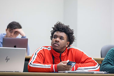 A student in a red jacket sits at a desk. Their phone is in their left hand and they are looking up and to the left.