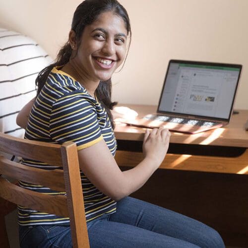 A student sits at a desk in their dorm room. They are wearing a black and white striped shirt and blue jeans. Their hands are resting on an open laptop.
