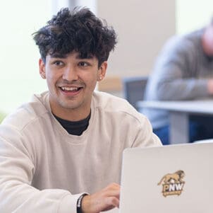 A student in an off-white sweatshirt sits at a table. There is a laptop open on the table.