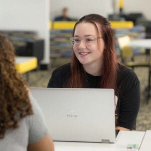 A student sits at a table with a laptop open in front of them. They are looking at another student off camera.