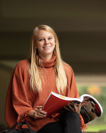 A student in an orange sweater holds an open textbook