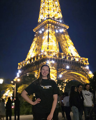 A student in an "I'm Powering Onward" t-shirt stands in front of the Eiffel Tower