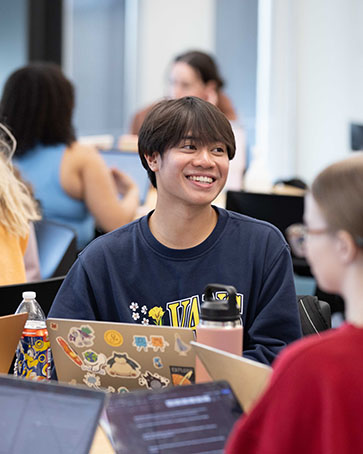 A student in a classroom. There are open laptops on the table around them.