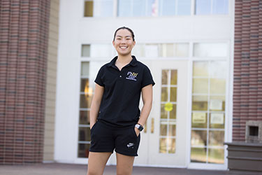 A student in a black PNW polo and black shorts stands outside of one of the university village buildings