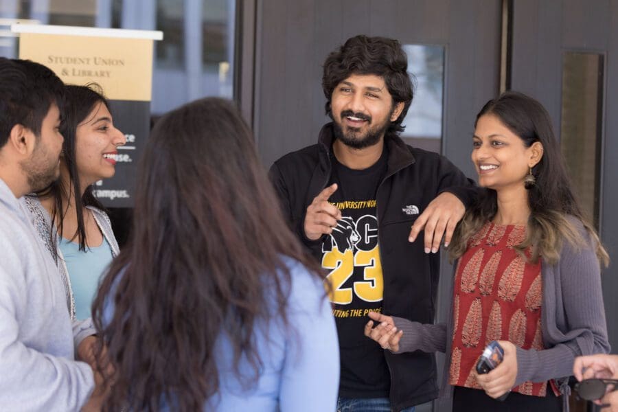 Students stand together outside. Two students are facing the camera and smiling. Three students have their backs to the camera