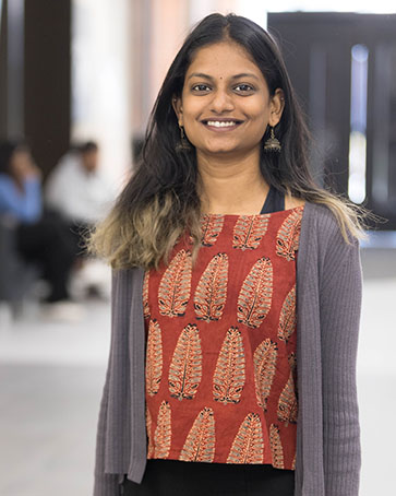 A student stands inside and smiles. They are wearing a red patterned shirt with a gray cardigan