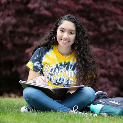 A student sits cross legged in the grass. They have a notebook open on their lap.