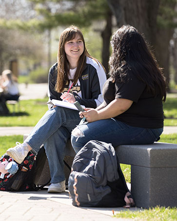 Two students sit on a concrete bench outdoors. They are turned toward each other