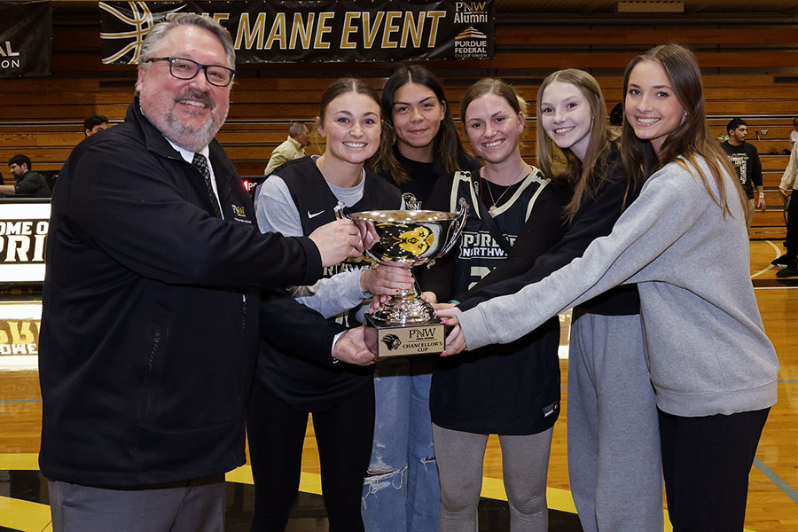 A man in a PNW jacket poses with a group of women holding a Chancellor's Cup trophy in front of a banner that says Mane Event.
