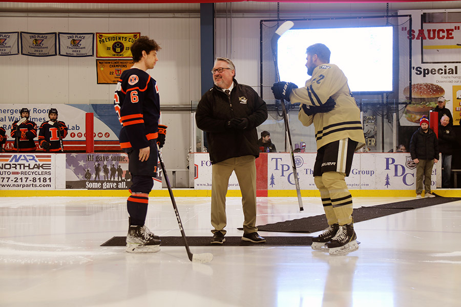 A man in a PNW jacket stands on the ice between two hockey players.