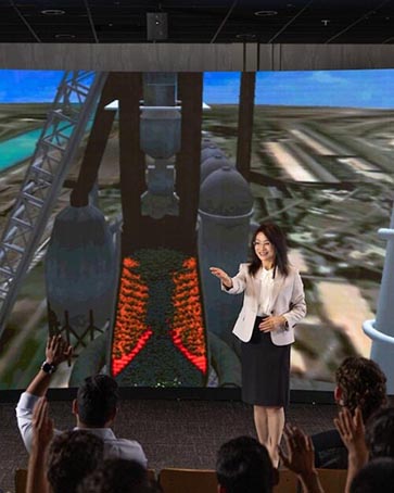 A woman stands in front of a visualization of a steelmaking blast furnace at Purdue University Northwest.