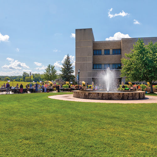 People gather in front of a building and fountain on PNW's Westville campus on a sunny day.