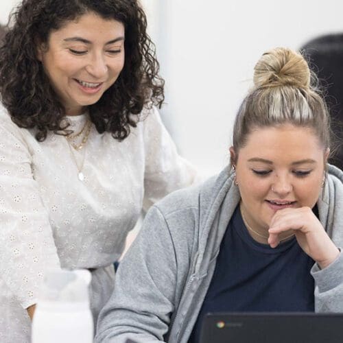 A student looks at a laptop. Their professor is standing behind them