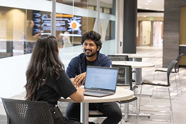 Two PNW students talking to each other in the Mane Zone while one student is working on their laptop 363x242
