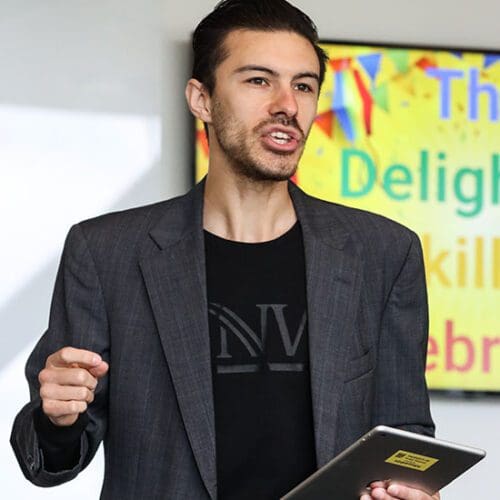 Nathan Elizalde stands with a tablet in his hand. There is a screen behind him that reads "The Delightful Skill of Celebration"