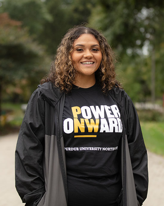 A student stands outdoors and smiles at the camera. They are wearing a "Power Onward" t-shirt and jacket.