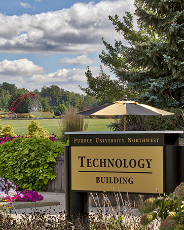 A sign on the Westville Campus reading "Purdue University Northwest Technology Building". The campus greenery and an umbrella are in the background.
