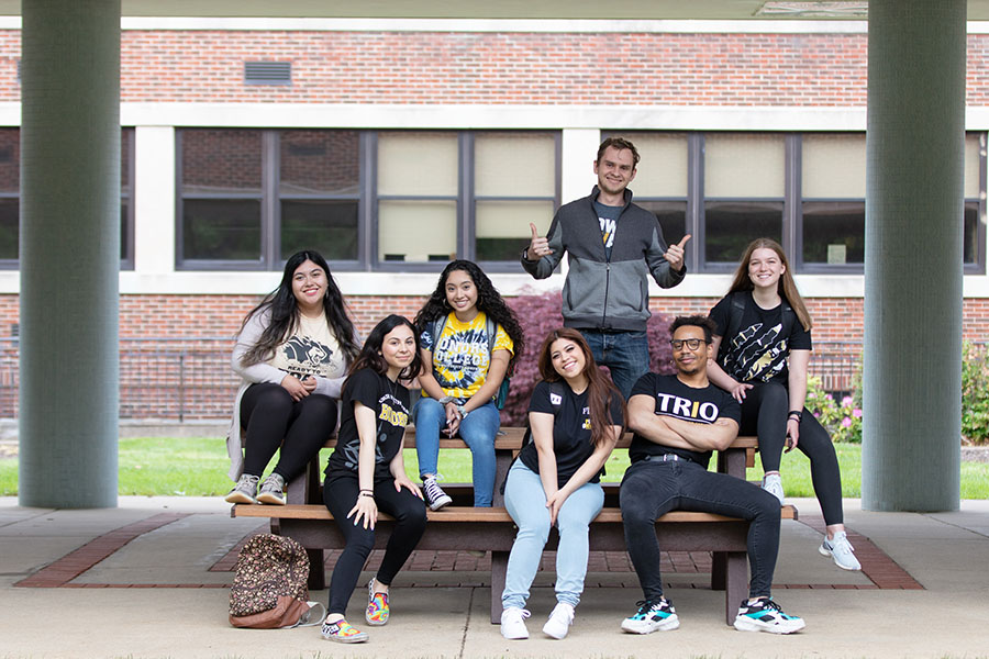 Seven students gather around a picnic table. One student is standing behind the table, the other six students are sitting in various positions on the table and bench