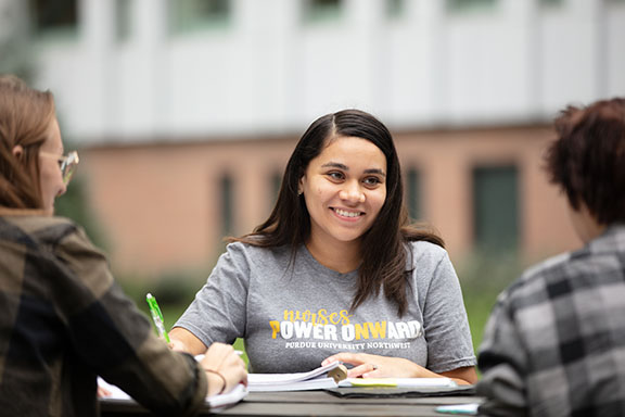 Student sits at a picnic table and smiles at the students sitting across from them.