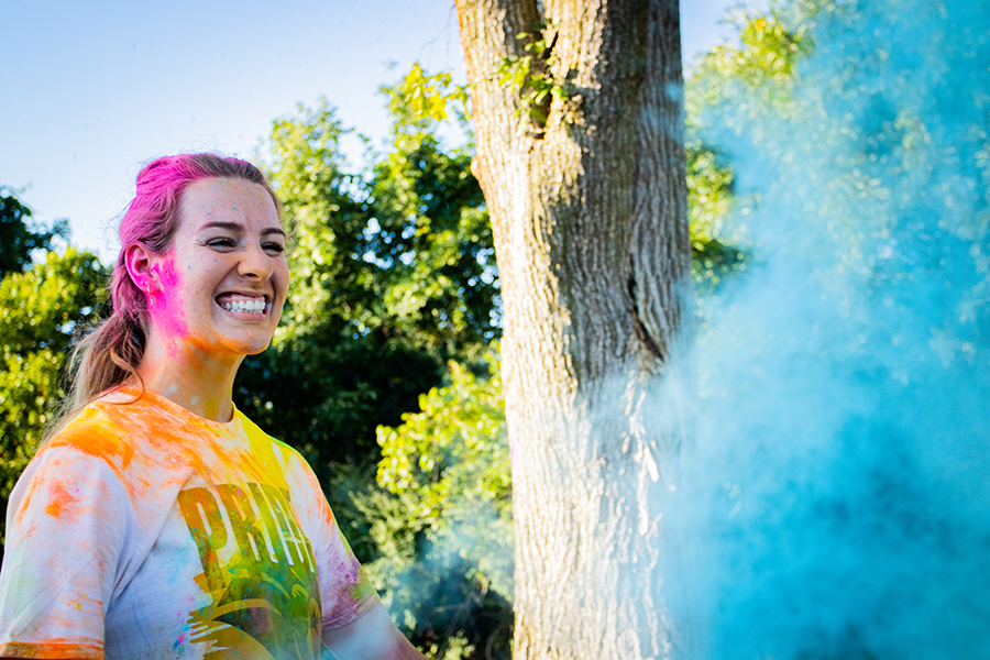 A student smiles while covered in color powder. There is a blue cloud of color powder in front of her.