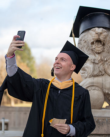 A student in commencement regalia takes a selfie in front of a lion sculpture wearing a cap and gown.