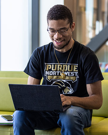 A student in a Purdue Northwest Athletics shirt works on a laptop on campus