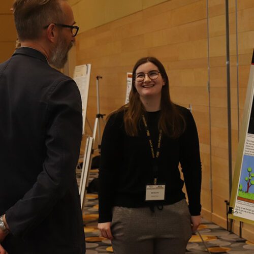 Two people stand near a printed research poster. They are looking at each other while talking.