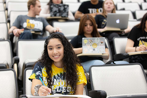 Students sit together in a tiered classroom. The student in the front is looking at the camera and smiling.