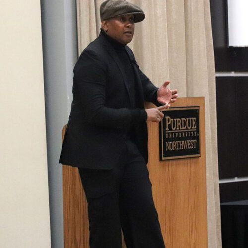 Kevin Powell stands on stage wearing a black quit and a gray hat. He is leaning against a podium