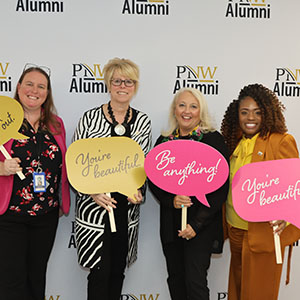 Four women stand together in front of a PNW alumni backdrop