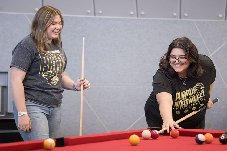 Two PNW students play pool on a red table in DSAC, both smiling and holding pool cues. They are wearing PNW shirts and jeans.