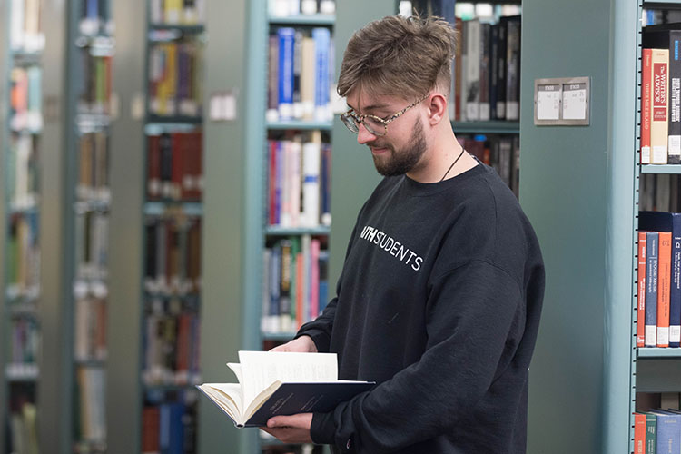 A PNW student in a black sweatshirt stands in the Hammond Campus library. The library stacks are pictured behind them.