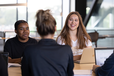 Three students talking together while working on their laptops in the NILS Building