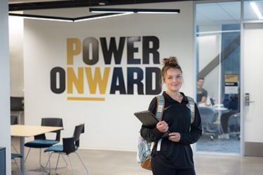 A student in a black PNW Athletics long sleeve poses while wearing a backpack and holding a laptop. Behind them on a white wall are the words "Power Onward".