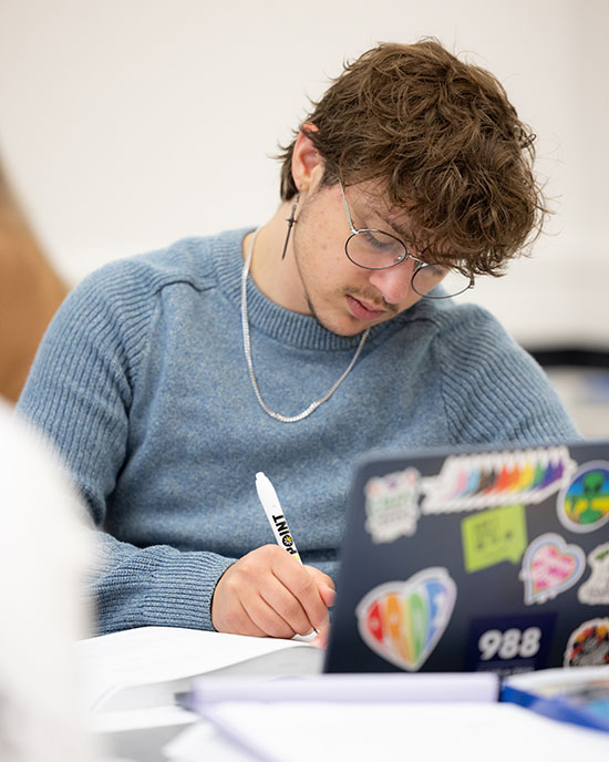 A PNW student sits at a desk and writes in a notebook. They have a laptop open on the desk in front of them.