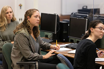 Students sit in front of computers. One student is taking notes on a piece of paper