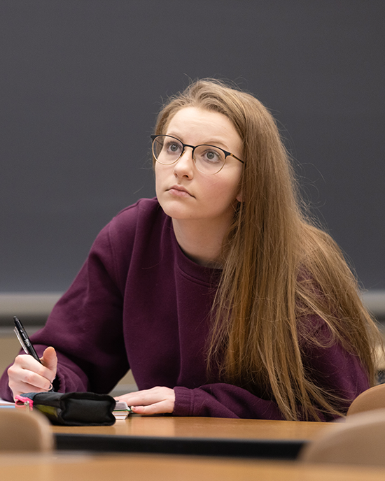 A student sits at a table. They are holding a pen in their right hand and resting their left hand on a notebook.
