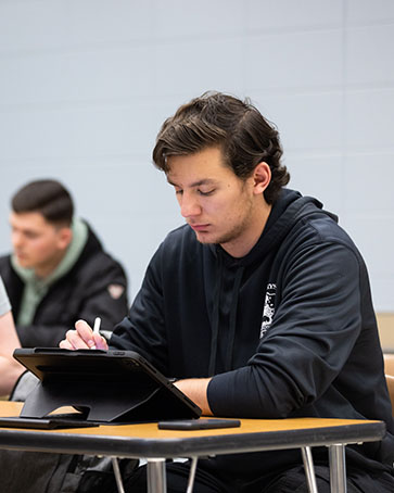 A student sits at a desk. They are writing on a tablet with a stylus pen