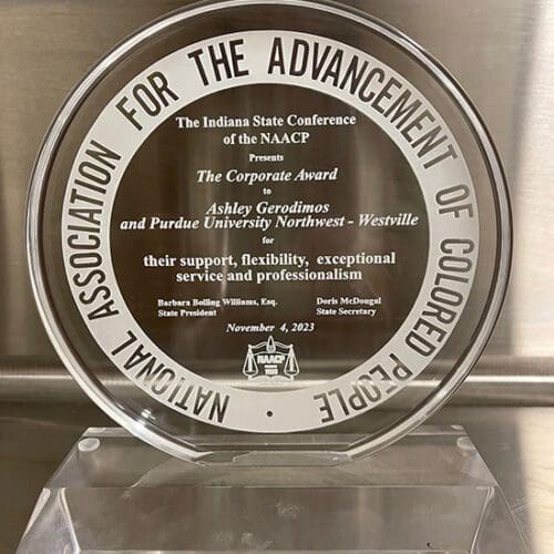 A circular glass award that reads "The Indiana State Conference of the NAACP presents The Corporate Award to Ashely Gerodimos and Purdue University Northwest - Westville for their support, flexibility, exceptional service and professionalism. Barbara Bolling Williams, Esq., State President. Doris McDougal, State Secretary. November 4, 2023. The NAACP logo is centered at the bottom of the award.