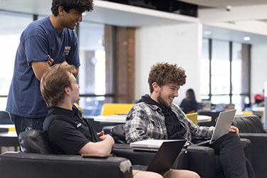 Two students sit in chairs with open laptops on their laps. One student is standing behind the chairs, leaning over and talking