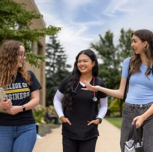 Three college students walking outdoors on campus and talking while carrying books and backpacks.
